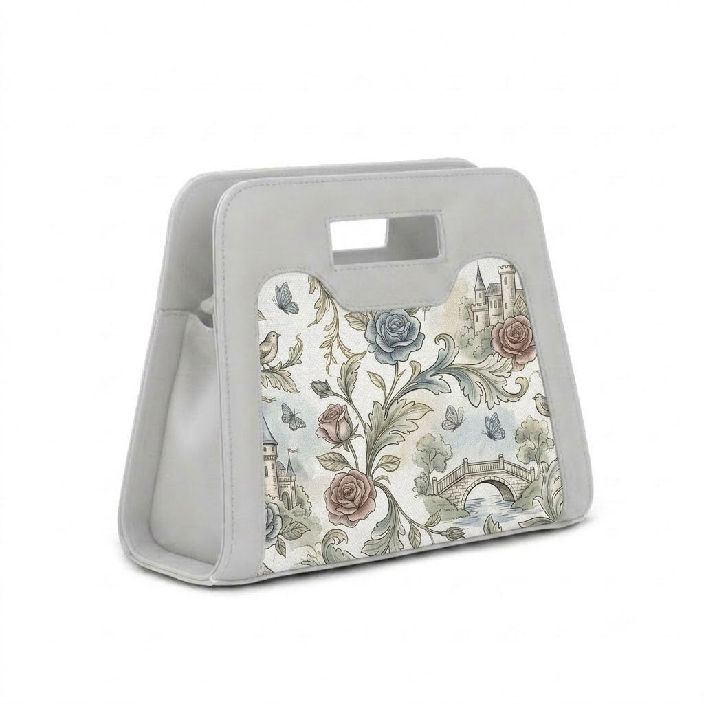 A modern, grey neoprene handbag with a cutout handle, featuring a detailed vintage print of castles, roses, and butterflies, positioned next to its white shoulder strap with black clasps against a white background.