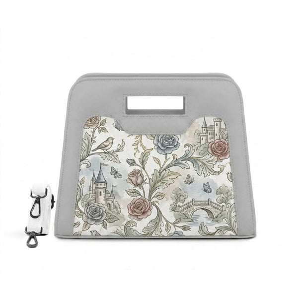 A modern, grey neoprene handbag with a cutout handle, featuring a detailed vintage print of castles, roses, and butterflies, positioned next to its white shoulder strap with black clasps against a white background.
